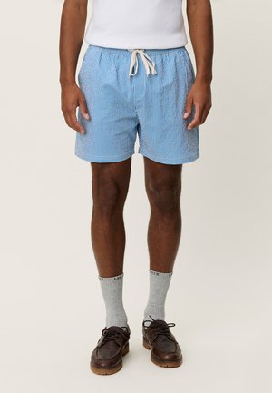 Modré a biele pruhované plavkové šortky s elastickým pásom, sťahovaním na šnúrku a voľným strihom, doplnené o sivé ponožky a hnedé topánky.