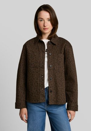 Jeune femme portant une veste marron à boutons imprimé léopard sur une chemise blanche et un jean bleu, debout devant un fond gris uni.