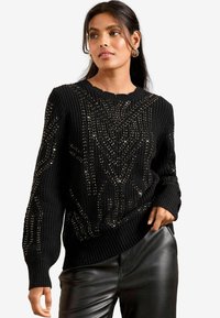 Schwarzer Strickpullover mit einem strukturierten Muster und metallischen Akzenten sowie einem gewellten Ausschnitt. Kombiniert mit Lederhosen.