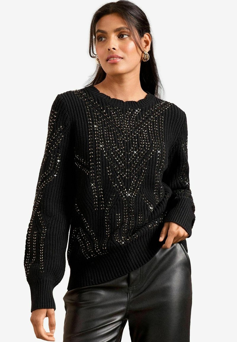 Schwarzer Strickpullover mit einem strukturierten Muster und metallischen Akzenten sowie einem gewellten Ausschnitt. Kombiniert mit Lederhosen.