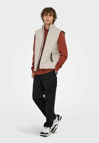 Hellbeige ärmellose Weste mit hohem Kragen, Frontreißverschluss und zwei Seitentaschen. Getragen über einem rostfarbenen Langarmshirt und schwarzen Hosen.