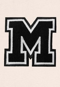 Lettera "M" ricamata in nero con contorno bianco su uno sfondo chiaro, caratterizzata da una finitura in tessuto testurizzato.