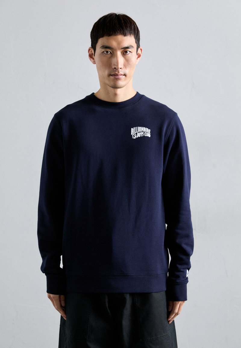Mann som har på seg en marineblå sweatshirts med "Billionaire Boys Club"-logoen på venstre bryst, står foran en enkel lys grå bakgrunn.