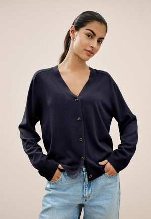 Marineblauwe cardigan van zacht materiaal, met een V-hals, knoopsluiting, verlaagde schouders en geribbelde mouwen. Gecombineerd met lichtblauwe jeans.
