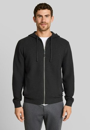Mann trägt einen dunklen Kapuzenpullover mit Reißverschluss und graue Hose, steht vor einem schlichten hellen Hintergrund und blickt leicht lächelnd nach vorne.