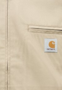 Béžová tkanina s horizontálnym rozopínateľným vreckom nad bielou štvorcovou náplasťou s oranžovým logom Carhartt a hnedým textom.