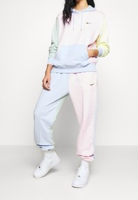Completo di felpa e pantaloni della tuta Nike in colori pastello, con un mix di pannelli rosa chiaro, blu, verde e giallo, arricchito da un accento del logo dorato.