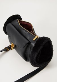 Borsa in pelle nera con apertura foderata in pelliccia, zip dorata e interno bordeaux; presenta una tracolla rimovibile e una base piatta.