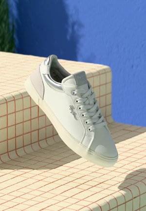 Sneaker grigio chiaro con tomaia in pelle liscia, dettagli metallici, design con lacci e suola in gomma testurizzata, posato su una superficie a motivi.
