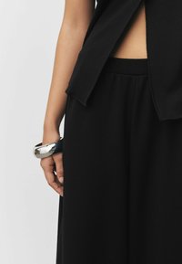 Schwarzes, locker sitzendes Oberteil mit seitlichem Schlitz, kombiniert mit schwarzen Hosen. Ein metallisches Armreif-ähnliches Armband am Handgelenk setzt einen Kontrast.