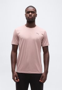 Άνδρας που στέκεται με το πρόσωπο προς τα εμπρός, φοράει ανοιχτό ροζ μπλουζάκι Puma και μαύρο παντελόνι, μπροστά σε απλό ανοιχτό φόντο.