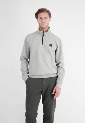 Mann steht mit den Händen in den Taschen, trägt hellgraues Viertel-Zip-Sweatshirt und dunkelgraue Hose vor weißem Hintergrund.