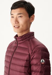 Chaqueta acolchada burdeos con cuello alto, cierre de cremallera y distintiva placa del logo de JOTT en el hombro izquierdo. Textura suave y ligera.