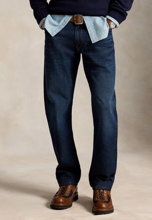 Mand iført mørkeblå jeans, lyseblå skjorte uden i bukserne, brun bælte, marineblå sweater og brune lædersko, står med hænderne i lommerne.