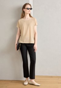 Beige t-shirt met korte mouwen, zwarte op maat gemaakte broek en lichtgekleurde platte schoenen. Model houdt een zwarte clutch vast en draagt een zonnebril.