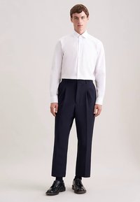 Weißes Hemd mit Knopfleiste kombiniert mit hoch taillierter, plissierter dunkelblauer Hose, ergänzt durch schwarze Lederschuhe. Glatte Texturen, maßgeschneiderte Passform.