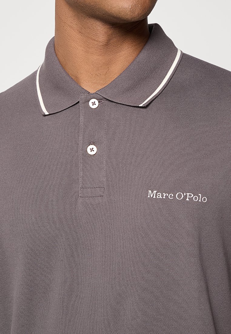 Szary golf z teksturowanej tkaniny, z białymi lamówkami na kołnierzyku oraz małym haftowanym logo po lewej stronie. Dwie guziki z przodu.