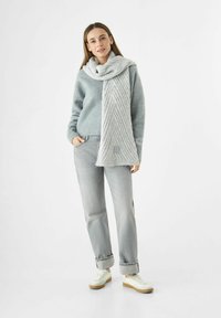 Vrouw in een grijze trui, lichte grijze jeans, witte sneakers en een dikke, lange grijze sjaal om haar nek, tegen een witte achtergrond.