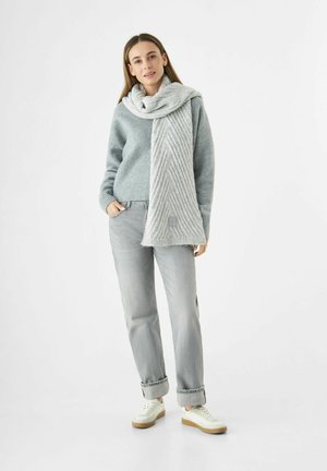 Vrouw in een grijze trui, lichte grijze jeans, witte sneakers en een dikke, lange grijze sjaal om haar nek, tegen een witte achtergrond.