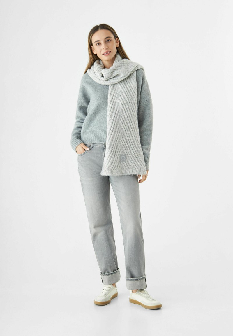 Vrouw in een grijze trui, lichte grijze jeans, witte sneakers en een dikke, lange grijze sjaal om haar nek, tegen een witte achtergrond.