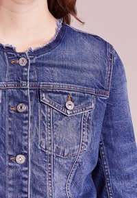 Jeansjacka i mellanblått med fransig krage, silverknappar och två bröstfickor med synlig sömdetalj och en texturerad tyg.