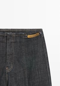 Ciemnoniebieskie dżinsy o prostym kroju, z widocznym pasem i złotymi elementami na froncie. Obecne są również przeszycia.