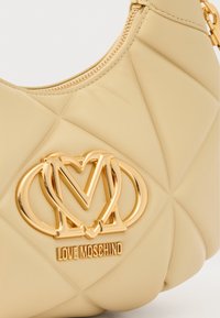 Μπεζ καπιτονέ τσάντα χειρός, με λογότυπο και φερμουάρ σε χρυσή απόχρωση. Μαλακό υλικό με μοτίβο διαμαντιού στο ράψιμο.