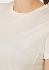 Hvid atletisk skjorte med rund halsudskæring. Har netpaneler på siderne. Subtil branding med teksten "ENDURANCE" i lys skrifttype.