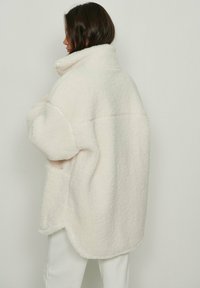 Chaqueta de forro polar blanco con un cuello alto, ajuste oversize y dobladillo redondeado. Textura suave con costuras cosidas, que presenta mangas cortas.