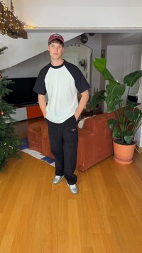 Zwart-wit T-shirt met korte mouwen, zwarte broek en zilveren schoenen. Staand in een kamer met een plant en een bruine bank.