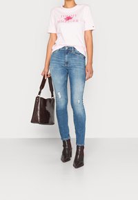 Camiseta de manga corta rosa claro con texto "Tommy Hilfiger", combinada con jeans ajustados azul medio. Bolso de mano efecto cocodrilo marrón oscuro y botines.