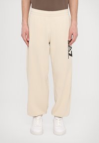 Beige sweatpants with an elastic waistband, wide legs, and a black logo detail on the side. Worn with white sneakers.
