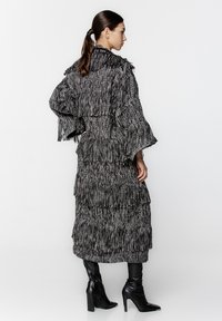Czarny i biały frędzlowany długi płaszcz o teksturowanym, warstwowym wzorze. Posiada szerokie rękawy i wysoki kołnierz, zestawiony z czarnymi botkami sięgającymi do kolan.
