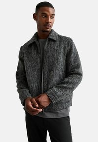 Chaqueta gris texturizada con cierre de cremallera, cuello y puños acanalados. Llevada sobre una camisa oscura, cuenta con un ajuste casual y un diseño moderno.