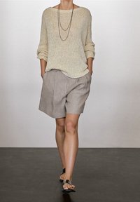 Kobieta spacerująca w beżowym swetrze z dzianiny, taupe plisowanych szortach, czarnych sandałach i warstwowych naszyjnikach na jednolitym tle.