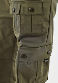 Groene tactische jackdetail met meerdere flapzakken, drukknoopsluitingen en een gladde, duurzame stof met een semi-gestructureerde pasvorm.