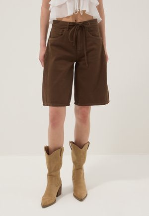 Mujer con pantalones cortos marrones hasta la rodilla con cintura ajustable, una camiseta blanca recortada con volantes y botas de ante marrón claro hasta media pantorrilla.