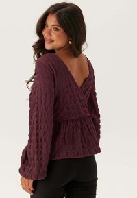 Bordeaux lange mouwen top met een gestructureerde wafelweef, v-back ontwerp en peplum taille. Glad materiaal en een losse pasvorm accentueren de vorm.