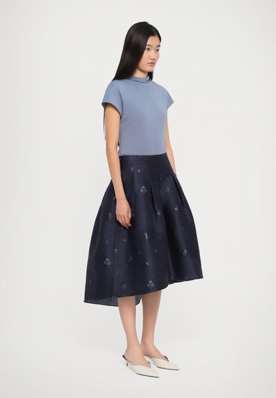 Woman with long black hair wearing a blue short-sleeve top, navy pleated midi skirt with subtle floral patterns, and white pointed heels.