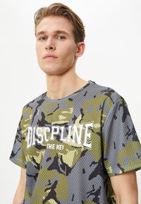 Camiseta de malla camuflada con cuello redondo y un atrevido texto blanco que dice "LA DISCIPLINA ES LA CLAVE". Los colores incluyen patrones en verde, gris y negro.