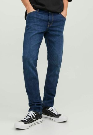 Jeans in denim blu scuro con una vestibilità slim, dotati di cinque tasche, cuciture sottili e indossati con sneakers nere e bianche.
