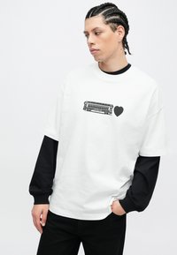 Maglietta bianca oversize con grafica nera stampata raffigurante un tram e un cuore. Indossata sopra una maglia a maniche lunghe nera. Tessuto in cotone.