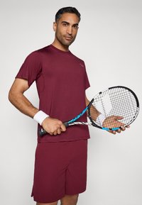 Burgundské športové tričko s krátkymi rukávmi a zodpovedajúce šortky. Drží čierno-modrý tenisový raket, na zápästí má biely náramok.