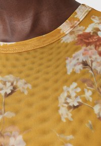 Geel nettop met een bloemenpatroon en witte en roze accenten. Ronde halslijn met een textuurafwerking en lichtdoorzichtig materiaal.
