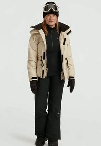 Mujer con chaqueta de esquí beige, pantalones de nieve negros, guantes, gorro negro y gafas de esquí en la frente, de pie frente a un fondo blanco.