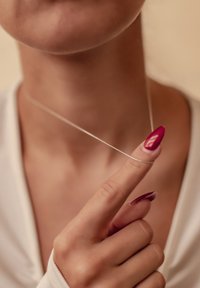Collar de plata sostenido por un dedo, con un diseño delgado y delicado. Esmalte de uñas rojo brillante en los dedos, fondo suave con tonos neutros.