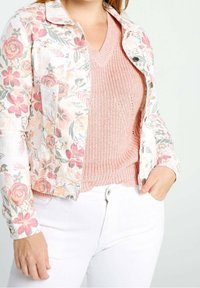 Giacca di jeans floreale con una morbida palette di colori pastello, caratterizzata da motivi rosa e verdi, abbinata a un maglione a V rosa chiaro in maglia e jeans bianchi.
