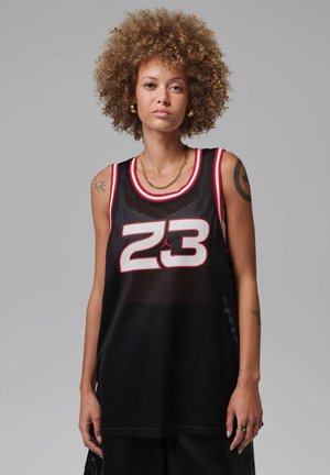 Persona joven con cabello rizado que lleva una camiseta deportiva negra con el número 23, collares dorados y tatuajes visibles en los brazos.