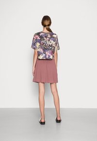 Haut à manches courtes à motif floral en gris foncé, associé à une jupe plissée couleur blush. Le mannequin porte des sandales plates noires. Fond épuré.