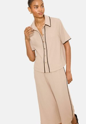 Frau trägt beige Kurzarm-Hemd mit Knöpfen und weit geschnittene Hose mit schwarzem Besatz, posiert mit Hand nahe am Kragen und schaut zur Seite.